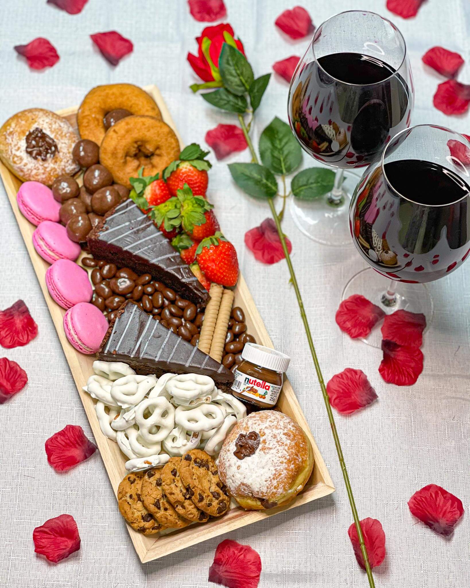 Cured catering valentine’s day dessert tray with donuts, macarons, cake and rose petals and wine